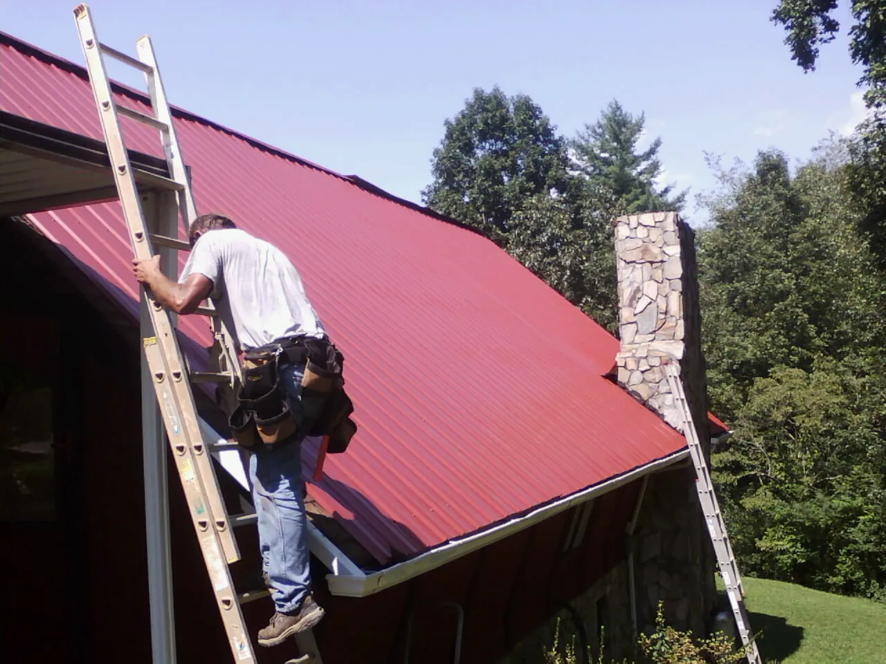 Roofer on a ladder beside a red metal roof and stone chimney — Soffit & Fascia Repair by Roofing Brothers in Hamilton City